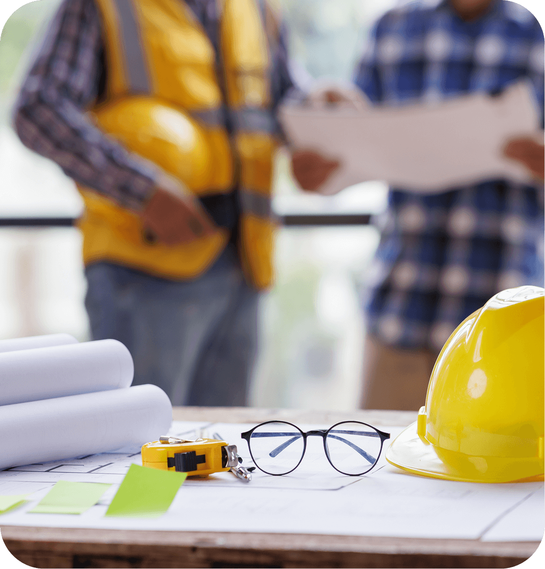 Close-up of a table with a yellow hard hat, eyeglasses, blueprints, and measuring tape. Two people wearing yellow safety vests and helmets are discussing papers in the background.